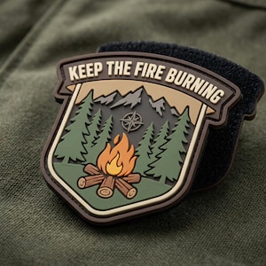 May include: A shield-shaped morale patch featuring a campfire, pine trees, mountains, and a compass rose. The text KEEP THE FIRE BURNING is printed in a tan banner at the top. The patch has a dark brown border and a black hook-and-loop backing.