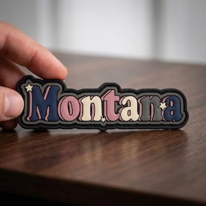 May include: A colorful Montana patch with a retro design. The word "Montana" is in a layered font with navy blue, pink, cream, and gray colors, outlined in black. Small star accents are included.
