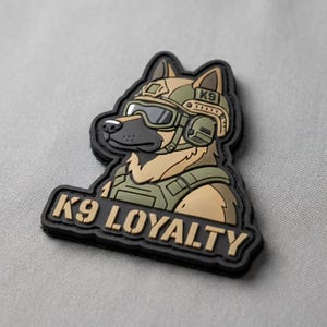 May include: A tactical morale patch featuring a cartoon German Shepherd dog wearing a tan helmet, tactical goggles, and a green vest. The dog has a black muzzle and tan fur. Below the dog, the text K9 LOYALTY is embossed in tan letters on a black background.