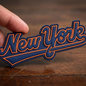 May include: A navy blue and orange New York emblem. The word "New York" is written in a stylized script with an orange outline. The emblem is held by a hand against a wooden background.