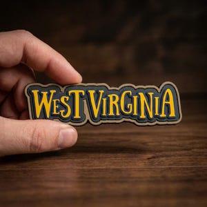 May include: A rectangular patch with the words "WEST VIRGINIA" in yellow, outlined in black and tan. The patch is held between fingers against a blurred wooden background. The text is in a bold, stylized font.
