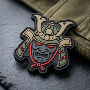 May include: A black, green, and tan patch featuring a stylized samurai helmet and mask design. The patch has red accents for the eyes and mouth, and is set against a textured background.