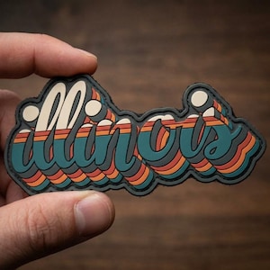 May include: A retro-style patch featuring the word "Illinois" in a layered, stylized font. The design incorporates teal, orange, and cream colors with a black border. The patch is held by a hand against a wooden background.