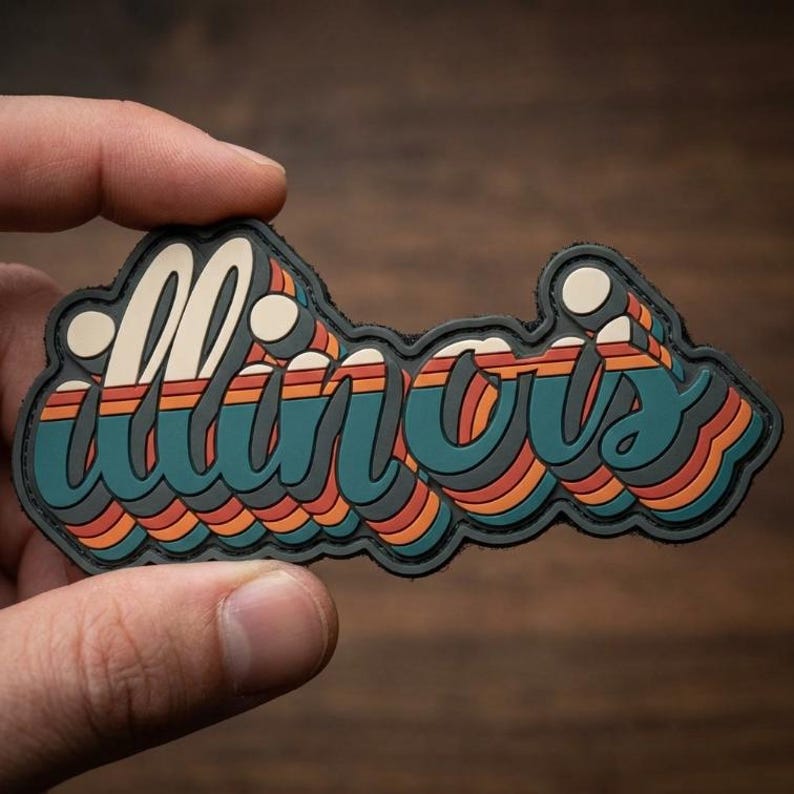 以下が含まれることがあります： レトロスタイルのパッチで、レイヤー状のスタイリッシュなフォントで「Illinois」の文字が描かれています。デザインは、ティール、オレンジ、クリーム色を使い、黒い縁取りが施されています。パッチは、木製の背景に対して手で持たれています。
