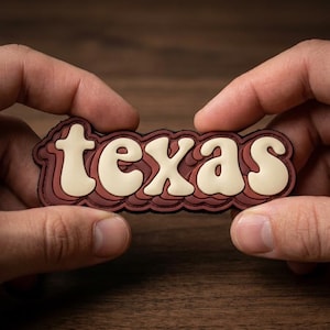 May include: A brown and cream-colored Texas patch with a layered design. The word "Texas" is in a cream-colored, retro-style font, outlined in brown, and set against a brown background.