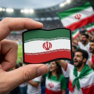 May include: A rectangular patch featuring the Iranian flag, held up in front of a crowd. The flag design includes green, white, and red horizontal stripes with a central emblem and Arabic script along the top and bottom edges.