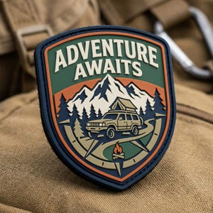 May include: A shield-shaped rubber patch featuring the text ADVENTURE AWAITS in cream letters. The design displays a cream-colored SUV with a roof tent, a campfire, pine trees, and snow-capped mountains against a green and orange background.