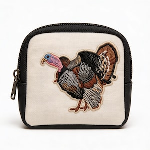 May include: A small, cream-colored zippered pouch with a detailed embroidered turkey design. The turkey features brown, gray, and black feathers, a pink and blue head, and is set against a cream background. The pouch has black leather trim and a black zipper.