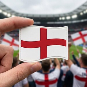 May include: A white and red England flag patch held between fingers, with a red cross on a white background. The background shows a stadium filled with people, some holding similar flags.