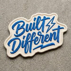 May include: Embroidered patch with the words "Built Different" in blue script and a lightning bolt design. The patch has a cream-colored background and a stitched border, set against a gray concrete surface.