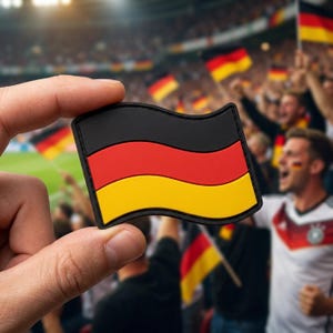 May include: A black, red, and yellow German flag patch is held up in front of a crowd of cheering fans. The patch has a black border and a wavy design. The background shows people waving flags and wearing German team apparel.