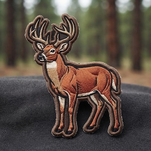 May include: An embroidered patch featuring a standing deer with large antlers. The deer has brown fur, a white neck and underbelly, and black hooves. The embroidery uses detailed stitching to define the animal's form and features against a dark border.