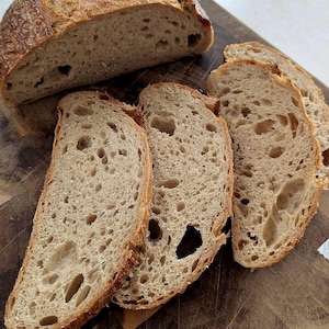 May include: A partially sliced loaf of bread and several slices on a wooden board. The bread has a golden-brown crust and a porous interior, indicating a sourdough or artisan style. The slices show air pockets. The bread is ready to eat.
