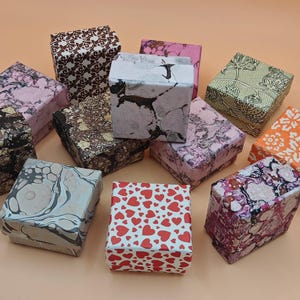 Può includere: Una collezione di dodici scatole regalo quadrate con vari motivi, tra cui cuori rossi, vortici marmorizzati in nero, bianco, rosa e grigio, stampe floreali in verde e arancione e disegni geometrici in marrone e bianco.