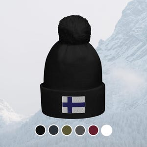 Puede incluir: Gorro negro con pompón, con la bandera de Finlandia bordada. La bandera presenta una cruz azul sobre fondo blanco. El gorro se muestra sobre un fondo de montañas nevadas. Disponible en varios colores.