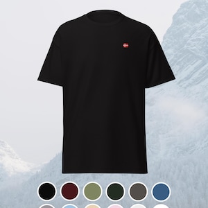 May include: Black t-shirt featuring a small embroidered Danish flag on the chest. The background shows a snowy mountain scene. A selection of potential shirt colours is displayed below the t-shirt.