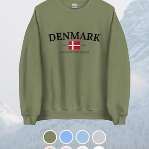 Könnte beinhalten: Olivgrünes Sweatshirt mit dem Wort "DENMARK" in Schwarz, einer dänischen Flagge und dem Text "EST 1965 LAND OF THE DANES". Das Sweatshirt hat einen Rundhalsausschnitt und eine Auswahl an Farboptionen unten. Der Hintergrund ist ein verschwommenes Bild eines Berges.
