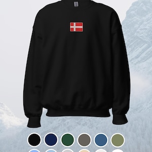 Könnte beinhalten: Schwarzes Sweatshirt mit Rundhalsausschnitt und kleinem, gesticktem dänischen Flaggenmotiv auf der Brust. Die Flagge zeigt ein weißes skandinavisches Kreuz auf rotem Grund. Das Sweatshirt hat gerippte Bündchen und einen gerippten Saum. Eine Farbpalette verschiedener Farbtöne wird darunter angezeigt.