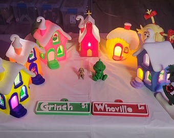 May include: A collection of illuminated miniature buildings and figures including a green character, a small dog, and two street signs labeled Grinch St and Whoville Ln. The colorful structures feature snow-dusted roofs and glowing windows in various hues.