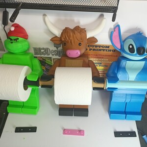May include: Three novelty toilet paper holders in the shape of cartoon characters. The Grinch, a Highland cow, and Stitch from Lilo & Stitch, each holding a roll of white toilet paper. The Grinch wears a red Santa hat. The cow has large white horns.