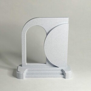 May include: A light gray, abstract sculpture with a speckled texture. The design features an arched opening, a semi-circular shape, and a base with three steps. The sculpture is a modern, minimalist art piece.