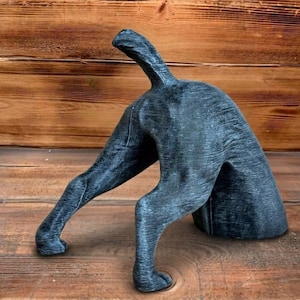 Può includere: Scultura di cane grigio scuro, raffigurante la parte posteriore di un cane con la coda sollevata e le zampe distese. La scultura è realizzata in un materiale strutturato e poggia su una superficie di legno.