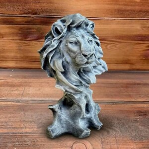 May include: A gray lion head sculpture with a detailed mane and facial features. The sculpture is mounted on a textured base, creating a regal and artistic appearance. The statue is made of a stone-like material.