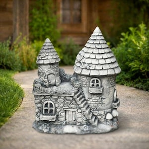 Peut inclure: Un ornement de jardin gris effet pierre en forme de petit château avec deux tours, un toit en bardeaux, des fenêtres arquées, une porte centrale et un escalier. La structure présente des détails de maçonnerie texturés et des vignes sculptées.