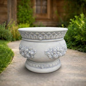 May include: A light grey stone planter featuring a wide, flared rim with a carved leaf pattern. The body of the planter is decorated with embossed clusters of roses and leaves, and it sits on a circular base with a small rose detail.