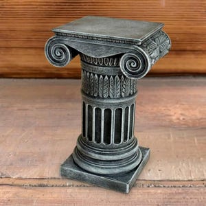 May include: A silver-colored, miniature Ionic column with intricate detailing. The column features a square base, a fluted shaft, and a decorative capital with scroll-like volutes. The overall design is reminiscent of classical architecture.