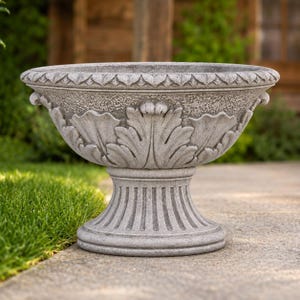 May include: A grey stone-effect pedestal planter featuring a wide, bowl-shaped top with a textured surface and carved acanthus leaf motifs. The base is fluted and circular, supporting the bowl, which has a decorative rim with a repeating pattern.