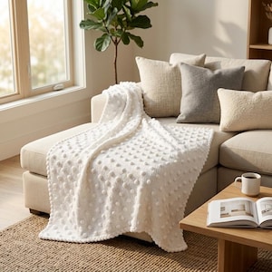 May include: A white textured throw blanket with a bobble design draped over a beige sofa. The sofa has several pillows in neutral tones. A wooden coffee table holds a mug and an open book. A window and a plant are in the background.