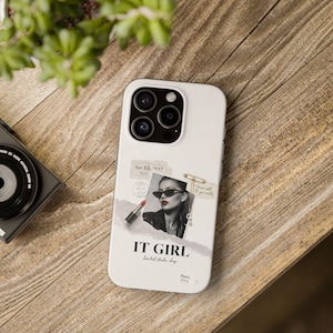以下が含まれることがあります： サングラスをかけた女性、口紅、そして「IT GIRL」の文字が特徴のコラージュデザインの白い携帯電話ケース。ケースにはトリプルレンズカメラの切り抜きがあり、木製の表面にあります。