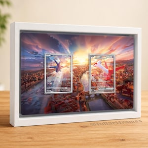 Pode incluir: Uma moldura retangular branca tipo caixa contendo duas cartas colecionáveis Pokémon, Latios e Latias, exibidas sobre uma impressão de paisagem urbana ao pôr do sol. A moldura apresenta um design limpo com um painel frontal transparente.