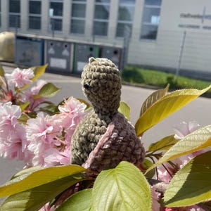 Puede incluir: Una figura de tortuga tejida a mano, de color verde oliva, con un acento rosa claro, ubicada entre flores de cerezo rosas y hojas verdes. La tortuga tiene un aspecto texturizado y un ojo pequeño y oscuro.