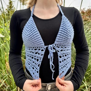 Op de afbeelding: Een lichtblauwe gehaakte haltertop met een driehoekig lijfje in een gestructureerd steekpatroon en een delicate kanten zoom. Het kledingstuk heeft dunne bandjes die in het midden aan de voorzijde worden gestrikt voor een open ontwerp.