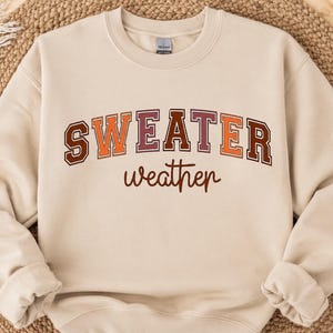 Peut inclure: Un sweat-shirt à col rond beige avec le texte "SWEATER" écrit dans une grande police universitaire arquée et multicolore avec des lettres marron, orange et bordeaux, suivi du mot "weather" dans une police cursive marron plus petite.