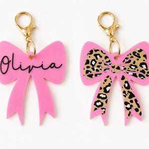 May include: Two pink bow-shaped keychains with gold-tone lobster claw clasps. The left keychain features the name Olivia in black script lettering. The right keychain displays a black and tan leopard print pattern across the surface of the bow.