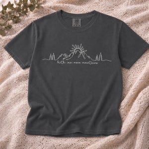 Könnte beinhalten: Dunkelgraues T-Shirt mit weißer Berg- und Sonnengrafik. Der Text "faith can move mountains" befindet sich darunter. Das Shirt besteht aus weichem Material und hat einen Rundhalsausschnitt.
