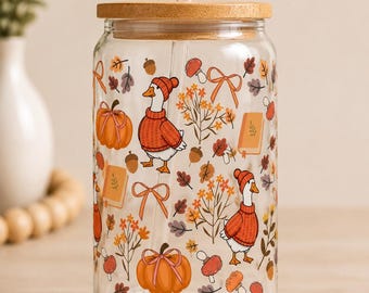 May include: A clear glass tumbler with a bamboo lid and a glass straw, featuring a repeating autumn-themed pattern of geese wearing orange sweaters and hats, pumpkins, acorns, mushrooms, books, and orange ribbons on a transparent surface.