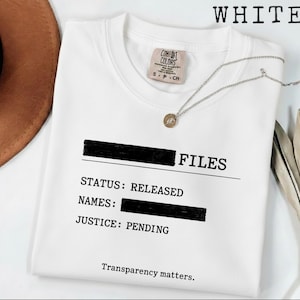 Könnte beinhalten: Weißes Sweatshirt mit Rundhalsausschnitt mit dem Wort „FILES“ und den Sätzen „STATUS: RELEASED“ und „JUSTICE: PENDING“ in Schwarz aufgedruckt. Der Text „Transparency matters.“ ist ebenfalls auf dem Shirt aufgedruckt. Eine silberne Halskette mit rundem Anhänger liegt auf dem Shirt.