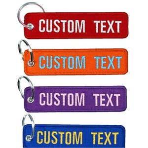 May include: Four rectangular keychains in red, orange, purple, and blue. Each keychain has the words "CUSTOM TEXT" embroidered in white or light blue, with a silver keyring attached.