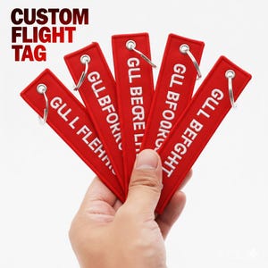 May include: A hand holding five red custom flight tags with white text and silver keyrings. The tags have different text, including "GLL FLEH", "GLL BFORK", "GLL BEQRE L", "GLL BFOOR", and "GLL BEFGHT". The words "CUSTOM FLIGHT TAG" are at the top.