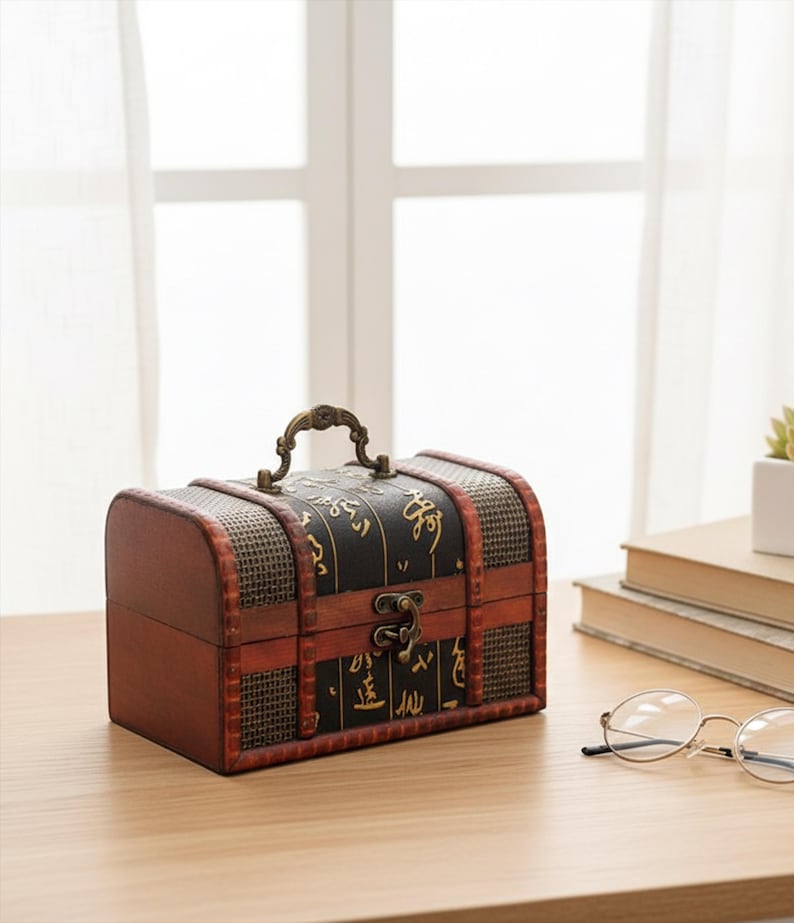 Op de afbeelding: Een kleine houten kist in antieke stijl met een gebogen bovenkant en een handvat. De kist is voornamelijk bruin met zwarte accenten en decoratief goudkleurig schrift. Het heeft een metalen sluiting en staat op een houten oppervlak.