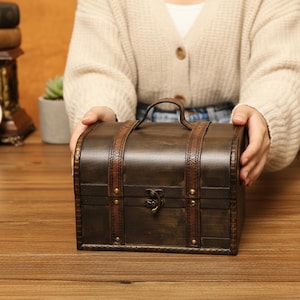 May include: A dark brown wooden chest with a curved top and a handle. The chest is accented with brown leather straps and a decorative metal clasp. The chest is held by a person.