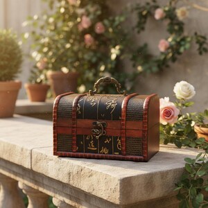 Op de afbeelding: Een kleine houten schatkist met een donkerbruine afwerking en decoratieve metalen accenten. De kist heeft een zwarte bovenkant met gouden kalligrafie en een messing handvat. De kist staat op een stenen richel met bloemen op de achtergrond.