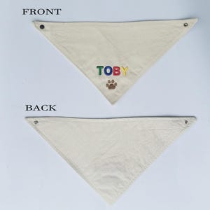May include: A triangular off-white pet bandana featuring the name TOBY embroidered in multi-coloured letters above a brown paw print icon. The bandana includes metal snap fasteners at the top corners and is labelled FRONT and BACK for clarity.