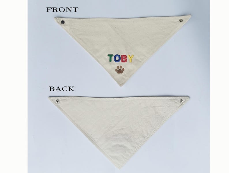 May include: A triangular off-white pet bandana featuring the name TOBY embroidered in multi-coloured letters above a brown paw print icon. The bandana includes metal snap fasteners at the top corners and is labelled FRONT and BACK for clarity.
