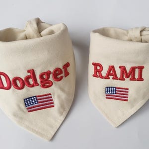 May include: Two off-white fabric pet bandanas featuring embroidered red text and a small American flag. The larger bandana displays the name Dodger, while the smaller bandana displays the name RAMI in red capital letters.