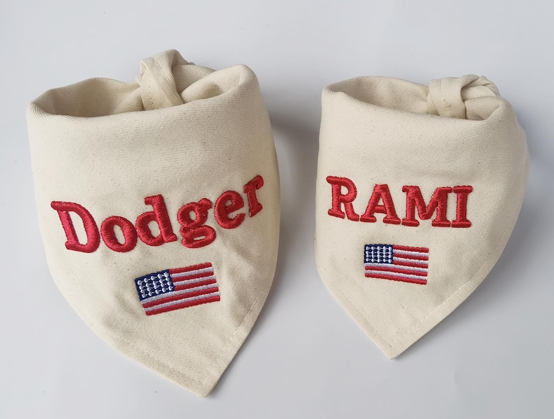 May include: Two off-white fabric pet bandanas featuring embroidered red text and a small American flag. The larger bandana displays the name Dodger, while the smaller bandana displays the name RAMI in red capital letters.